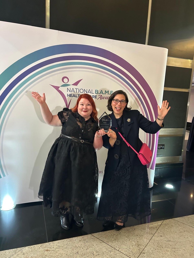 Two women stood in front of the BAME awards banner, holding their award